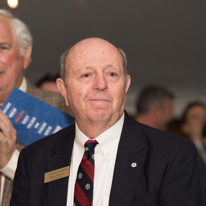 The image shows an older man with a bald head, wearing a dark suit, a white shirt, and a tie with red and black stripes. He has a name tag that says "Dick Finch". He is looking towards the camera. Another man is partially visible on the left side of the image.